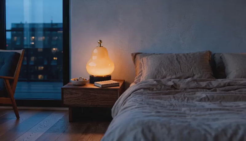 Cinematic bedroom video of a pear-shaped glass table lamp glowing warmly on a wooden nightstand beside linen bedding at dusk.
