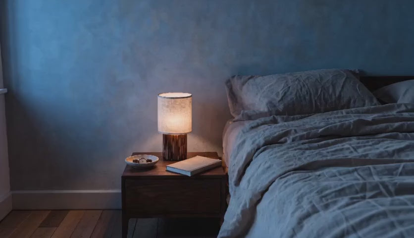 Charger la vidéo : Cinematic video of a cordless linen shade table lamp glowing on a wooden nightstand beside a linen-dressed bed at dusk.