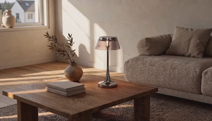 Charger la vidéo : Cinematic video of a cordless chrome table lamp with tinted acrylic drum shade glowing on a wooden coffee table beside a sofa.