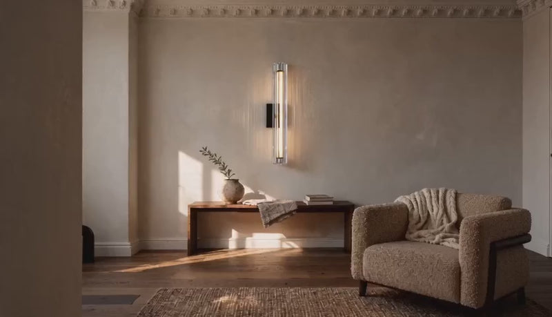 Cinematic video of a vertical fluted glass LED wall sconce glowing warmly above a wooden bench in a neutral living room.
