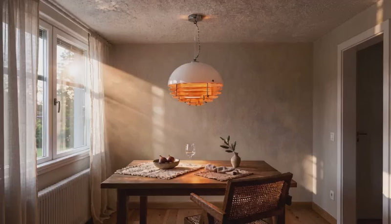 Cinematic video of a white glass dome pendant lamp with stacked orange acrylic rings glowing above a wooden dining table near a sunlit window.